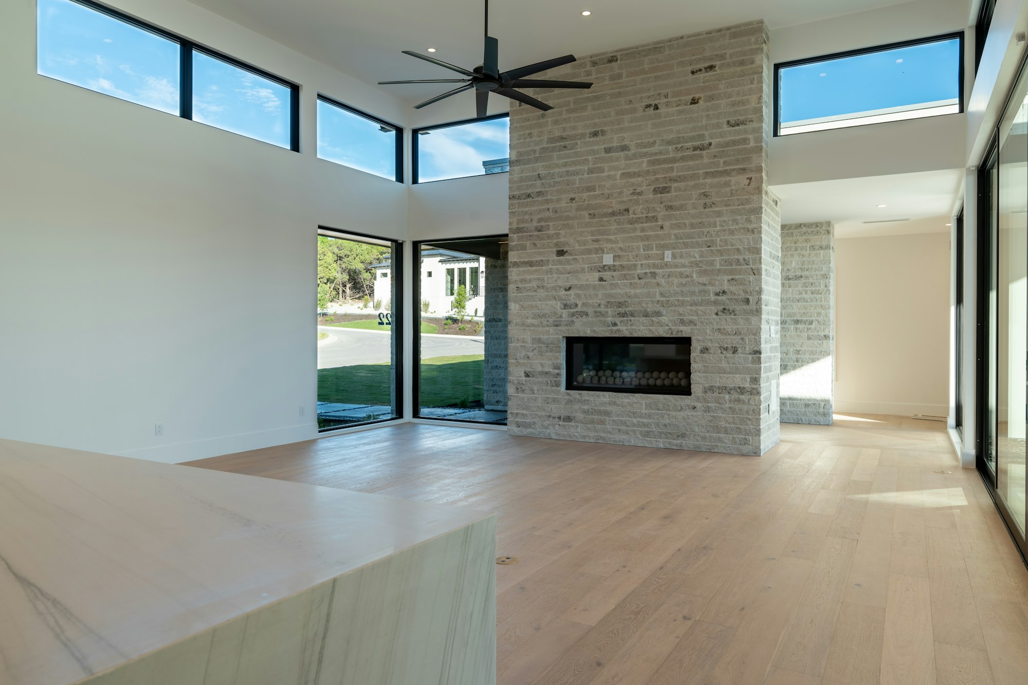Salón vacío con chimenea de ladrillo, suelo de madera clara y grandes ventanales